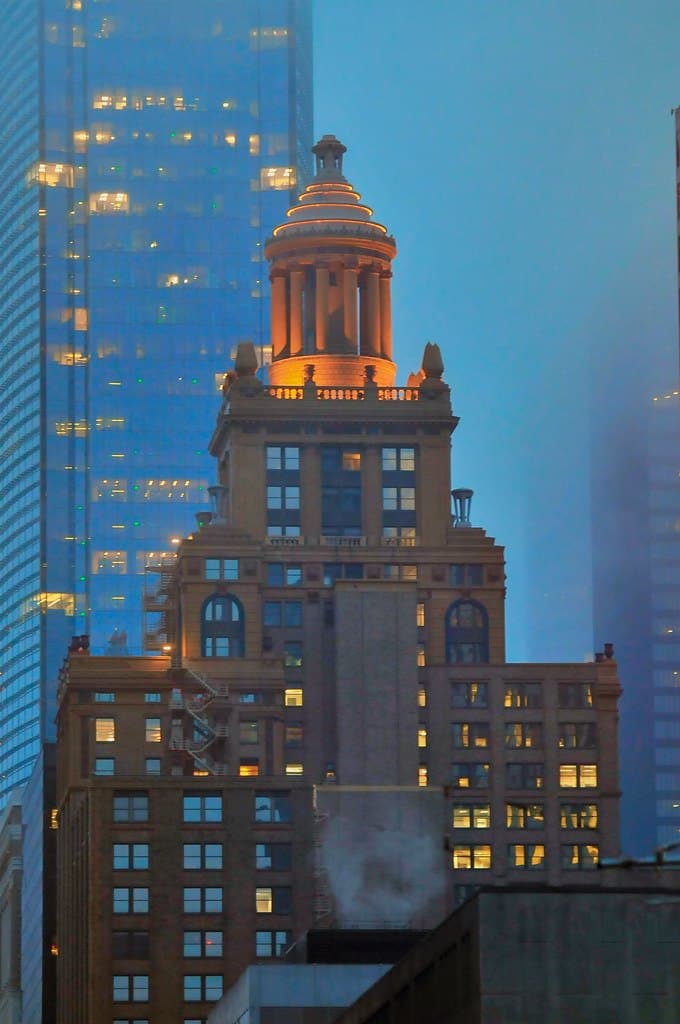 A historic stone office building with a lit cupola at dusk, in front of a glass skyscraper